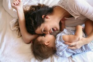 Moeder en kind die samen op bed liggen. De moeder lacht en spiegelt de lach van haar kind. Resonantie en responsiviteit zijn belangrijk om veilige hechting tot stand te brengen.